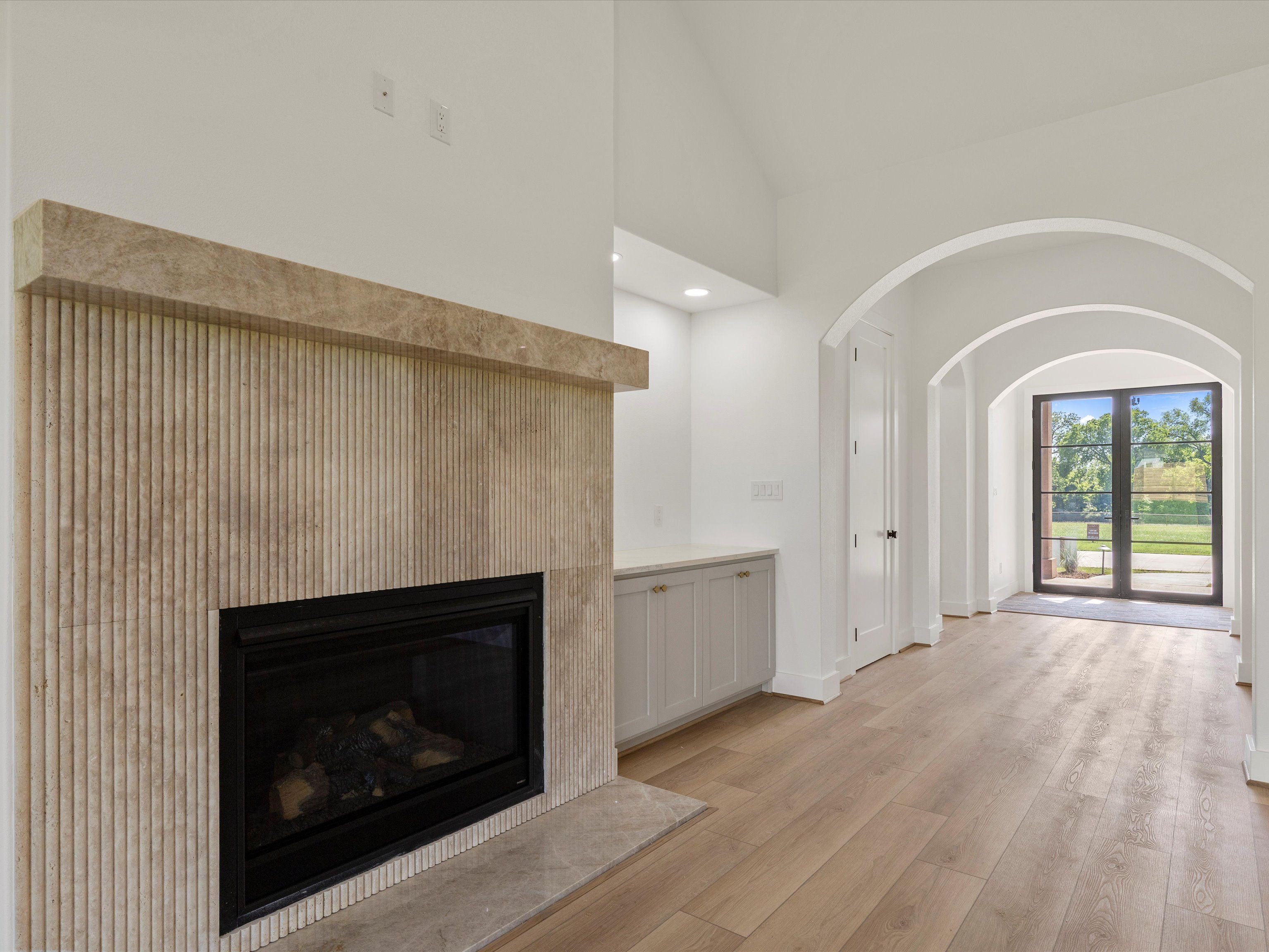 Interior view of a modern living space featuring a textured stone fireplace, light wood flooring, and arched doorways leading to an outdoor area.